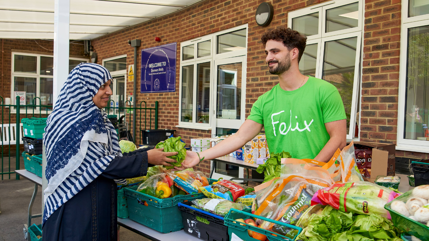 A man wearing a green t shirt featuring the logo of The Felix Project hands a lettuce to a visitor from a table full of colourful, healthy looking food items. They are both smiling