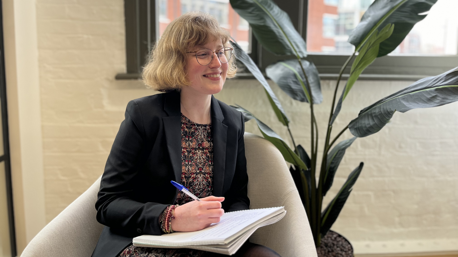 Head shot of Cicely Norman, joining Farrer Kane & Co as a PR Assistant. She sits in an office, writing in a notebook, with a green leafy plant in the background.